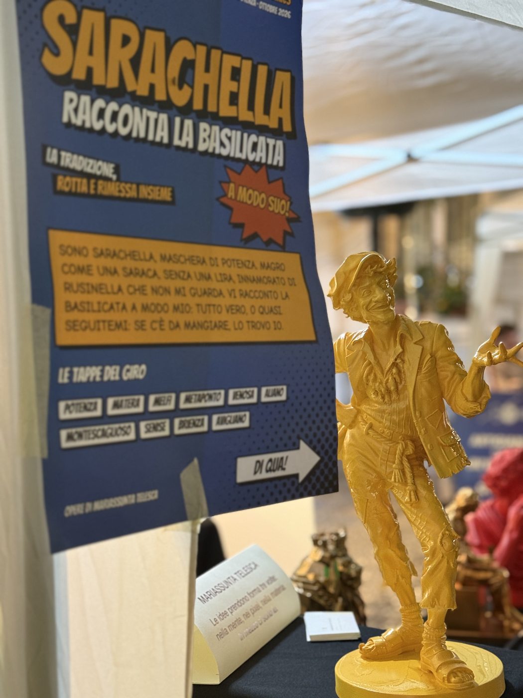 Sarachella, la maschera di Potenza, statua dorata accanto al manifesto «Sarachella racconta la Basilicata»